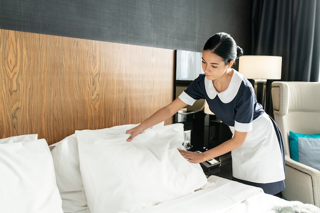 Молодая красивая горничная кладет подушку на белое чистое белье во время смены постели в одном из номеров отеля во время подготовки к новым гостям
