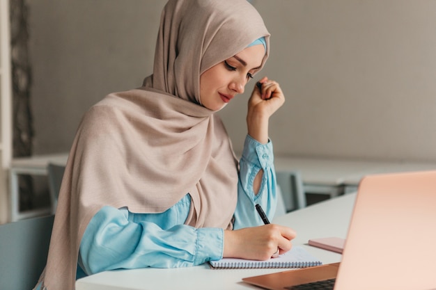 Young pretty modern muslim woman in hijab working on laptop in office room, education online