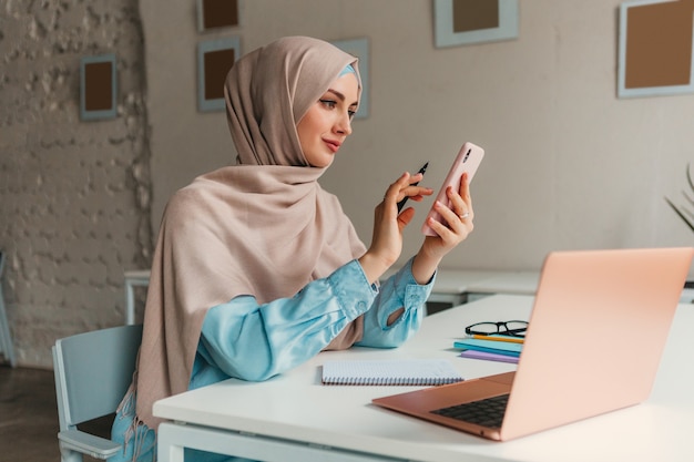 Young pretty modern muslim woman in hijab working on laptop in office room, education online