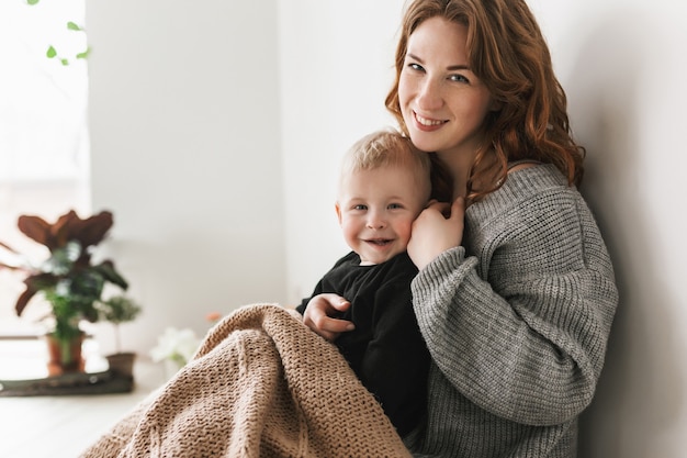 Young pretty mom with red hair in knitted sweater sitting on floor, holding her little handsome son