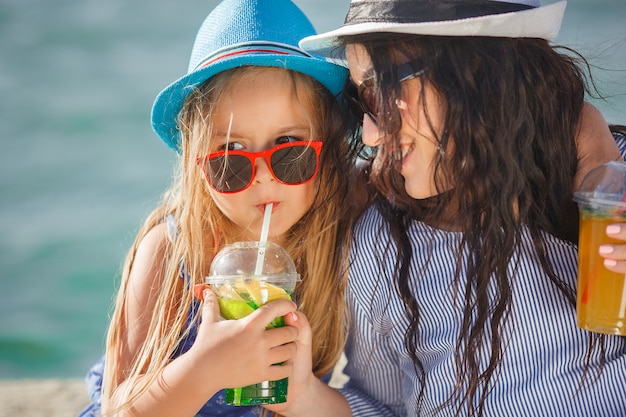 Young pretty mother and her little daughter on the beach having fun. Girls drinking lemonade.