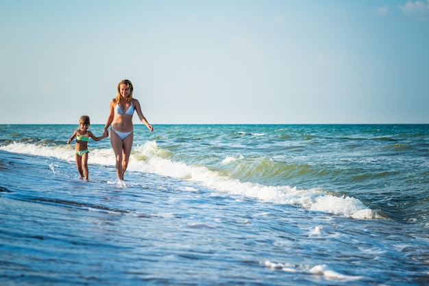 Giovane bella madre e figlia piccola corrono in riva al mare durante le vacanze in una soleggiata giornata estiva calda contro un cielo blu