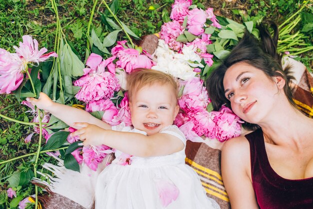 Young pretty mother with daughter lie on a plaid in the park. Family outdoor recreation. Family summer picnic in the park. Mother and daughter outdoors surrounded by pink peonies.