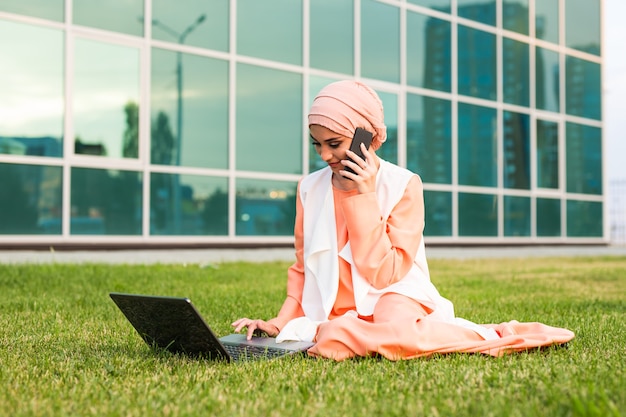 A young pretty muslim businesswoman in a park while talking on smartphone looking at the laptop