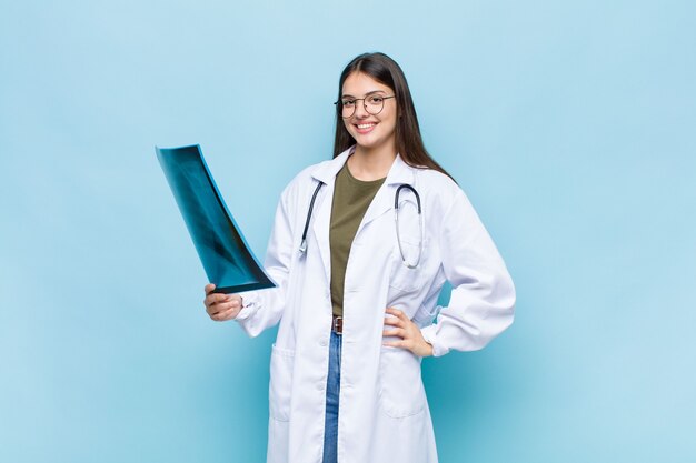 Photo young pretty physician smiling happily with a hand on hip and confident, positive, proud and friendly attitude