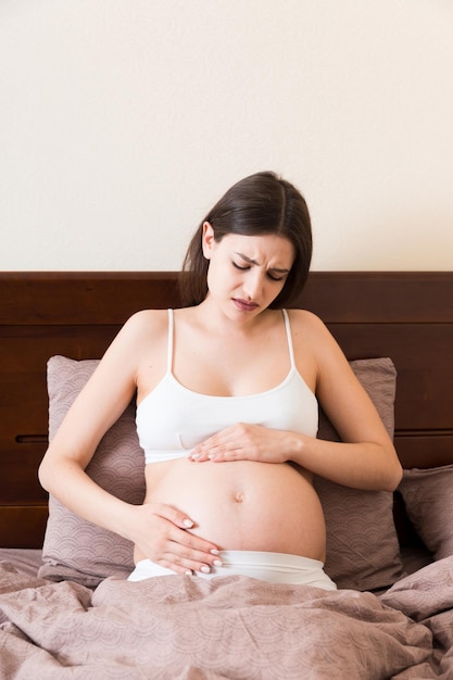 出産の準備をしている寝室のベッドに座って痛みを伴う若いかなり妊娠中の女性