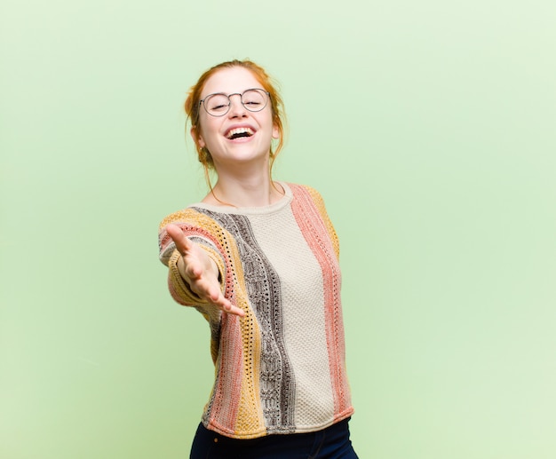 Young pretty red head woman smiling, looking happy, confident and friendly, offering a handshake to close a deal, cooperating against green wall