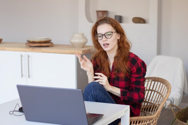 Giovane bella ragazza dai capelli rossi che parla usando un computer a casa in cucina