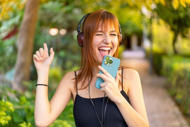 Young pretty redhead woman at outdoors listening music with a mobile and singing