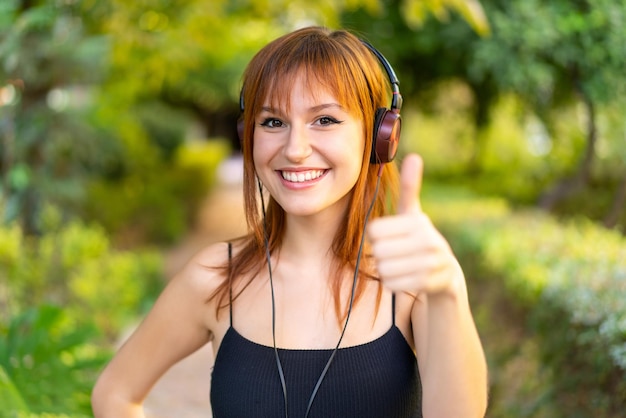 Young pretty redhead woman at outdoors listening music and with thumb up