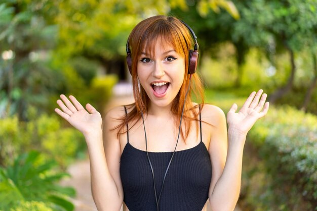 Молодая симпатичная рыжая женщина на улице удивлена и слушает музыку