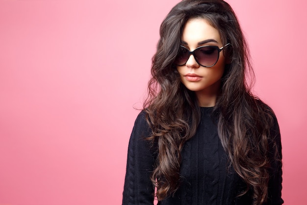 Young pretty sexy woman or girl with cute face and long brunette hair wearing sunglasses and black sweater, posing on pink background