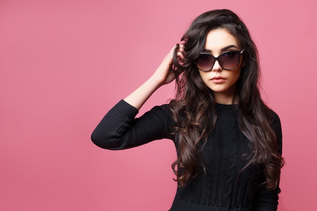 Young pretty sexy woman or girl with cute face and long brunette hair wearing sunglasses and black sweater, posing on pink background