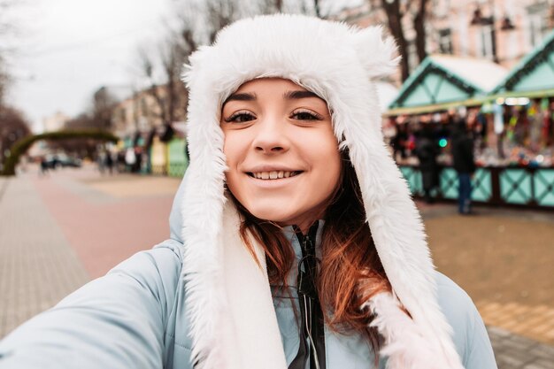 Young pretty smiling woman celebrating dressed warm jacket and woven woolen hat, taking sealfie