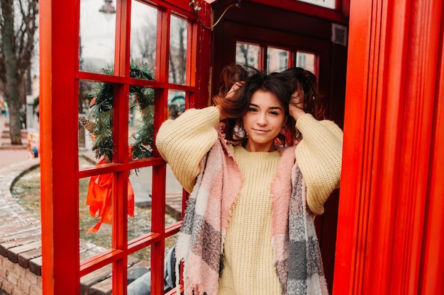 Young pretty smiling woman celebrating dressed warm sweater, standing near telephone red box