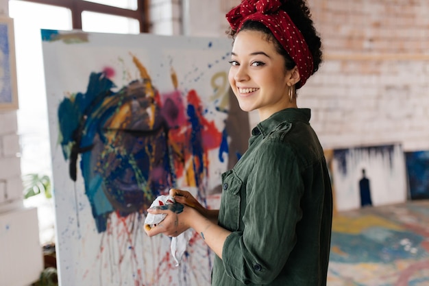 Foto giovane bella donna sorridente con capelli ricci scuri che disegna un'immagine su tela a mano con colori ad olio luminosi mentre guarda felicemente in camera in un grande laboratorio accogliente