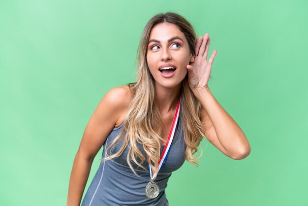 Young pretty sport Uruguayan woman with medals over isolated background listening to something by putting hand on the ear