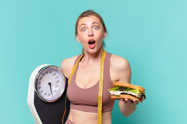 若いかわいいスポーツの女性は表情を驚かせ、体重計とサンドイッチを持っています