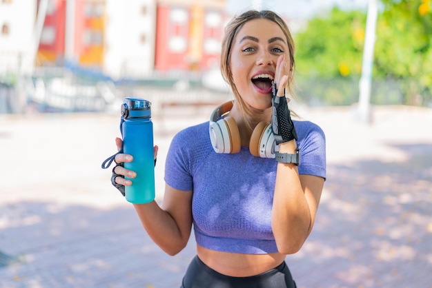 Young pretty sport woman with a bottle of water at outdoors shouting with mouth wide open