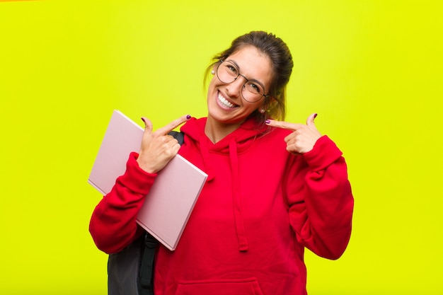 Young pretty student smiling confidently pointing to own broad smile, positive, relaxed, satisfied attitude