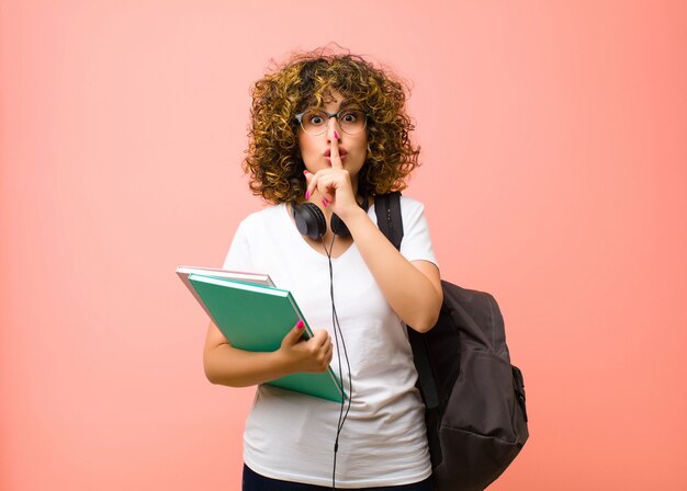 Foto giovane bella studentessa che chiede silenzio e tranquillità, gesticolando con un dito davanti alla bocca, dicendo shh o mantenendo un segreto contro il muro rosa