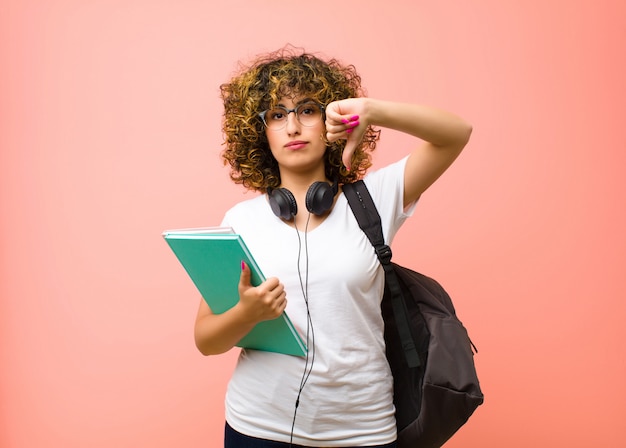 Young pretty student woman feeling confused, clueless and unsure, weighting the good and bad in different options or choices against pink wall