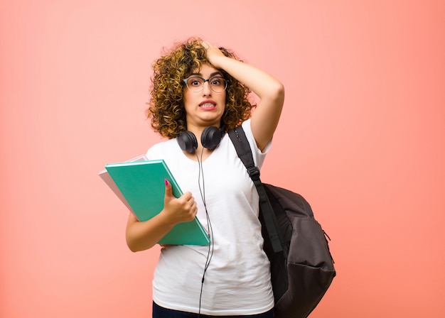 Foto giovane bella studentessa che solleva il palmo alla fronte pensando oops, dopo aver fatto uno stupido errore o aver ricordato, sentendosi muta contro il muro rosa