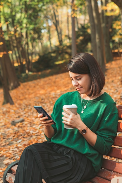 Young pretty stylish woman sitting in autumn park drinking coffee surfing internet on the phone. copy space