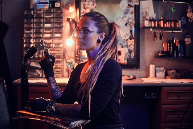 Young pretty tattoo master with dreadlocks and glasses is taking short break while holding tattoo machine.