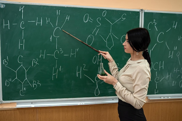 Young pretty teacher learn lesson on chemistry and standing near blackboard with chemical formula