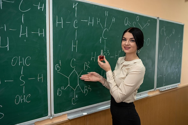 Young pretty teacher learn lesson on chemistry and standing near blackboard with chemical formula