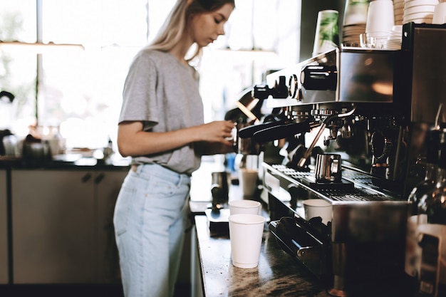 A young pretty thin blonde,dressed in casual outfit,is cooking coffee in a popular coffee shop. .