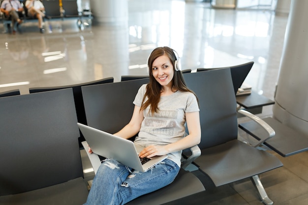 노트북에서 작동하는 음악을 듣고 헤드폰을 가진 젊은 예쁜 여행자 관광 여자, 국제 공항의 로비 홀에서 기다립니다