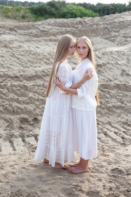 Молодые красивые близнецы с длинными светлыми волосами обнимаются в песчаном карьере в элегантном белом платье, юбке, куртке.