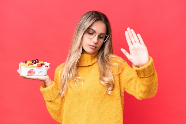 Foto giovane bella donna uruguaiana che tiene una ciotola di frutta isolata su sfondo rosso che fa gesto di arresto e delusa