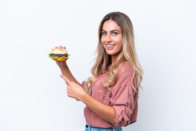 Молодая красивая уругвайская женщина держит бургер на белом фоне и указывает на него