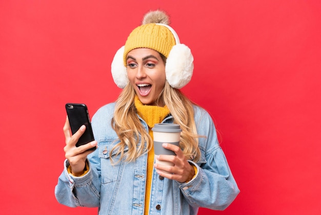 Young pretty Uruguayan woman wearing winter muffs isolated on red background background holding coffee to take away and a mobile