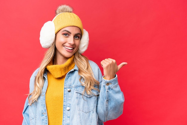 Young pretty Uruguayan woman wearing winter muffs isolated on red background background pointing to the side to present a product