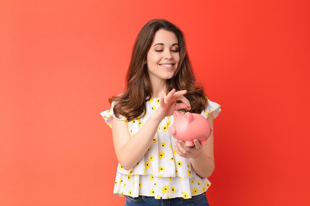 Young pretty woman against red wall holding a piggybank