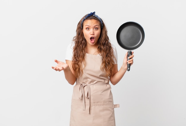 Young pretty woman amazed, shocked and astonished with an unbelievable surprise chef concept and holding a pan