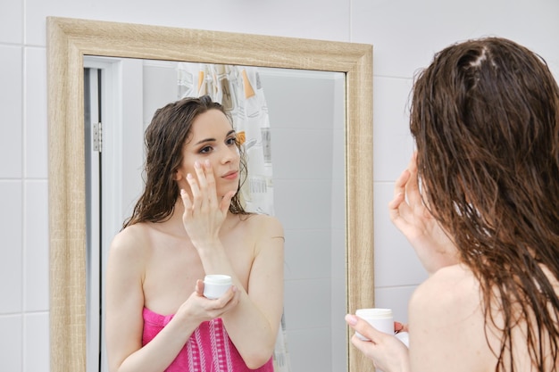 Young pretty woman applying cream on face skin while standing in front of mirror in bathroom