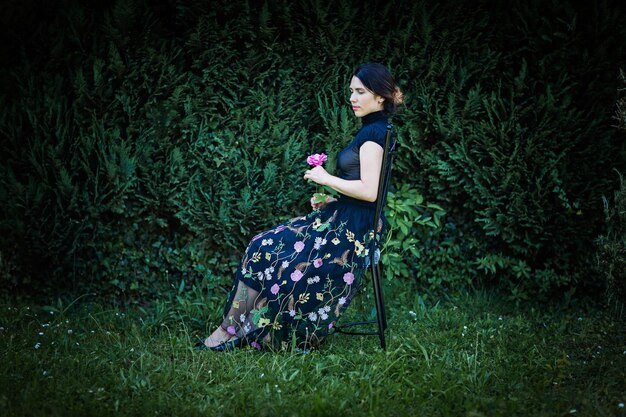 Young pretty woman in black clothes sits on a chair in the garden near a bush with a rose in her hands