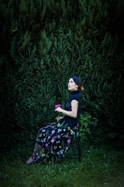 Young pretty woman in black clothes sits on a chair in the garden near a bush with a rose in her hands