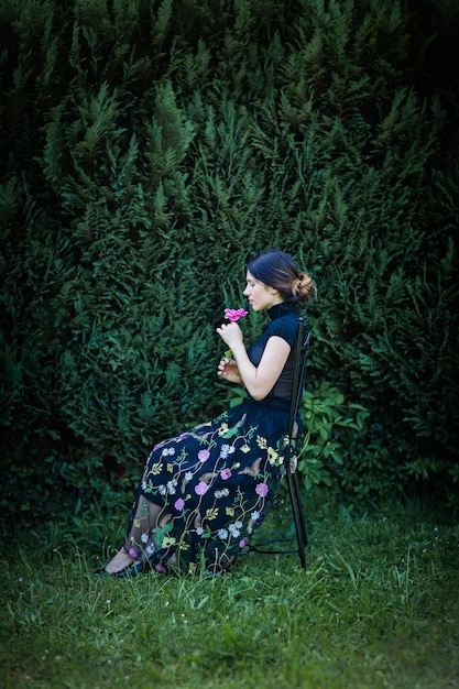 Young pretty woman in black clothes sits on a chair in the garden near a bush with a rose in her hands