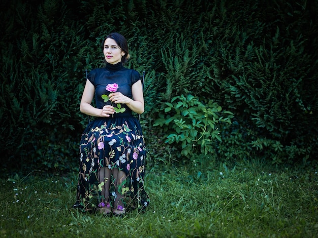 Young pretty woman in black clothes sits on a chair in the garden near a bush with a rose in her hands