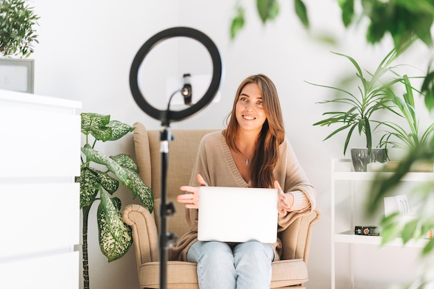 Young pretty woman blogger records video on mobile phone using ring lamp sitting on chair with laptop in room with green plants at home