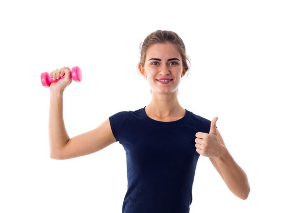 Young pretty woman in blue T-shirt holding pink dumbbell and showing thumb up on white background in studio