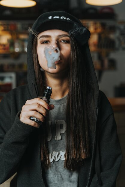 Foto giovane bella donna con il berretto fuma una sigaretta elettronica al negozio di vapori in primo piano in stile hiphop