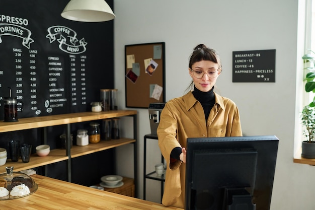 Young pretty woman in casualwear standing in front of computer by counter desk