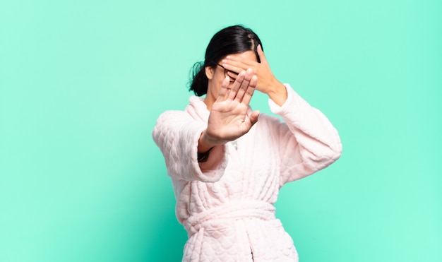 young pretty woman covering face with hand and putting other hand up front to stop camera, refusing photos or pictures. pajamas concept